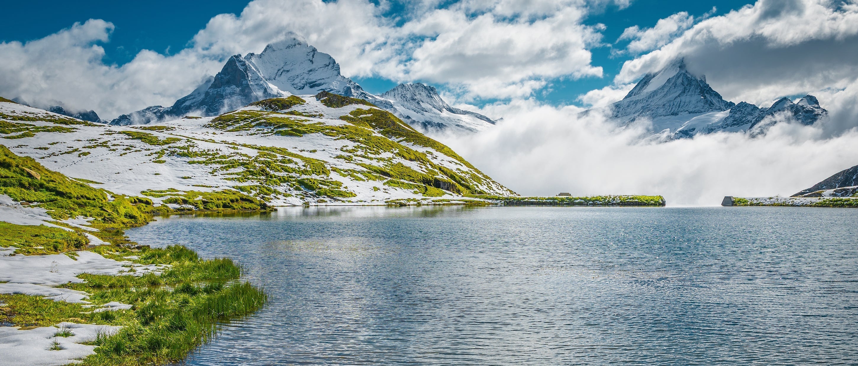 A scenic swiss mountain scernery with snow-dusted green slopes, a calm blue alpine lake, distant jagged peaks, and a partly cloudy sky.