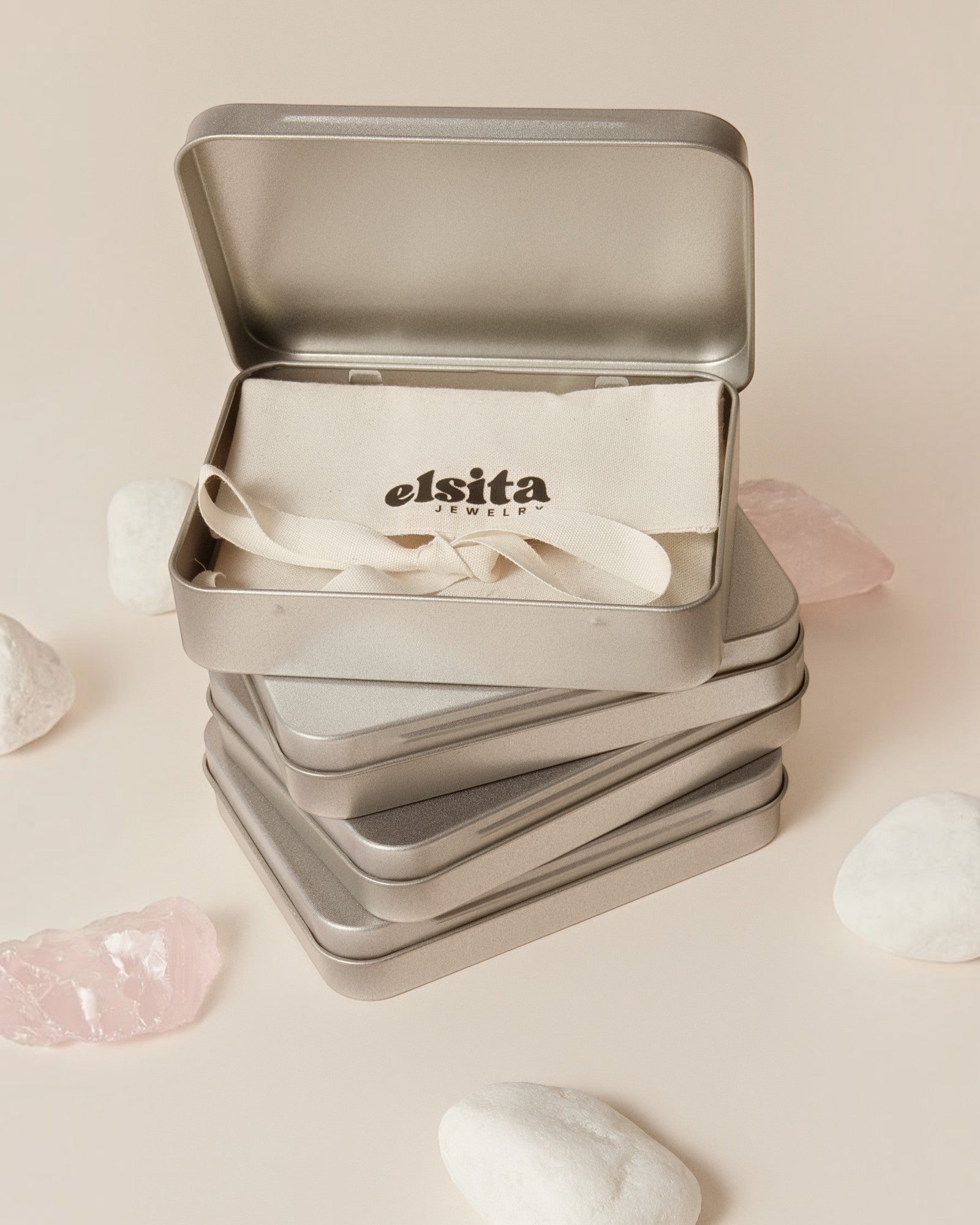 Elsita Jewelry branded metal gift tin containing a fabric pouch, surrounded by raw rose quartz stones on beige.