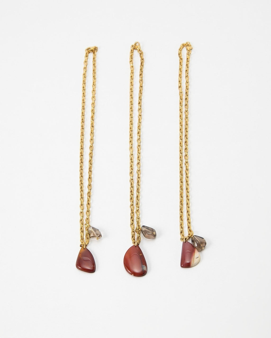 Three Elsita Jewelry Ember gold chain necklaces featuring teardrop-shaped red jasper and small smoky quartz charms.