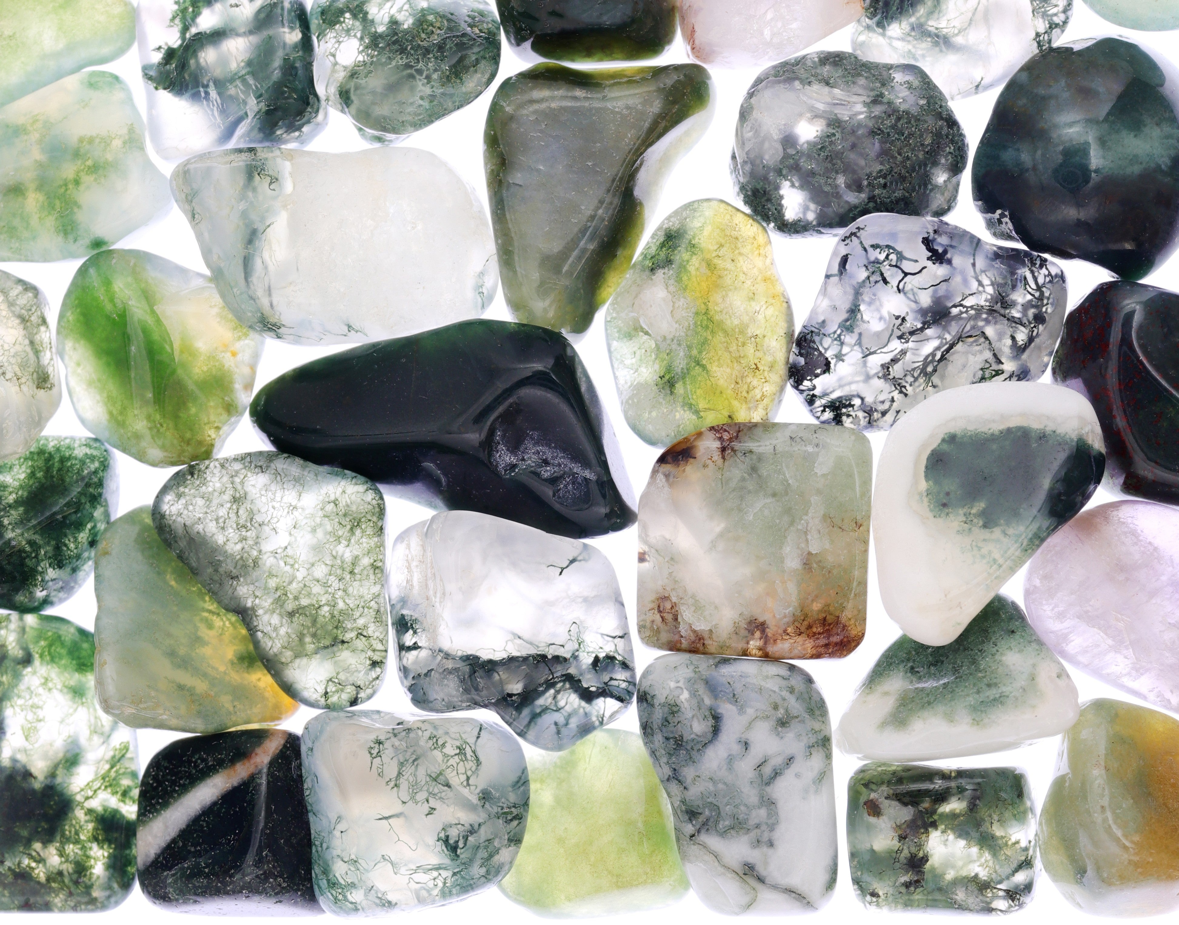 A collection of polished tumbled gemstones in various colors and patterns, including shades of green, white, black, gray, and hints of yellow and purple, arranged closely together on a white background.